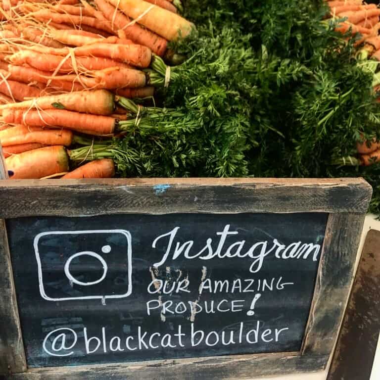 A blackboard at Black Cat Farm's stand with carrots on top and the Instagram account for the farm and restaurants @blackcatboulder